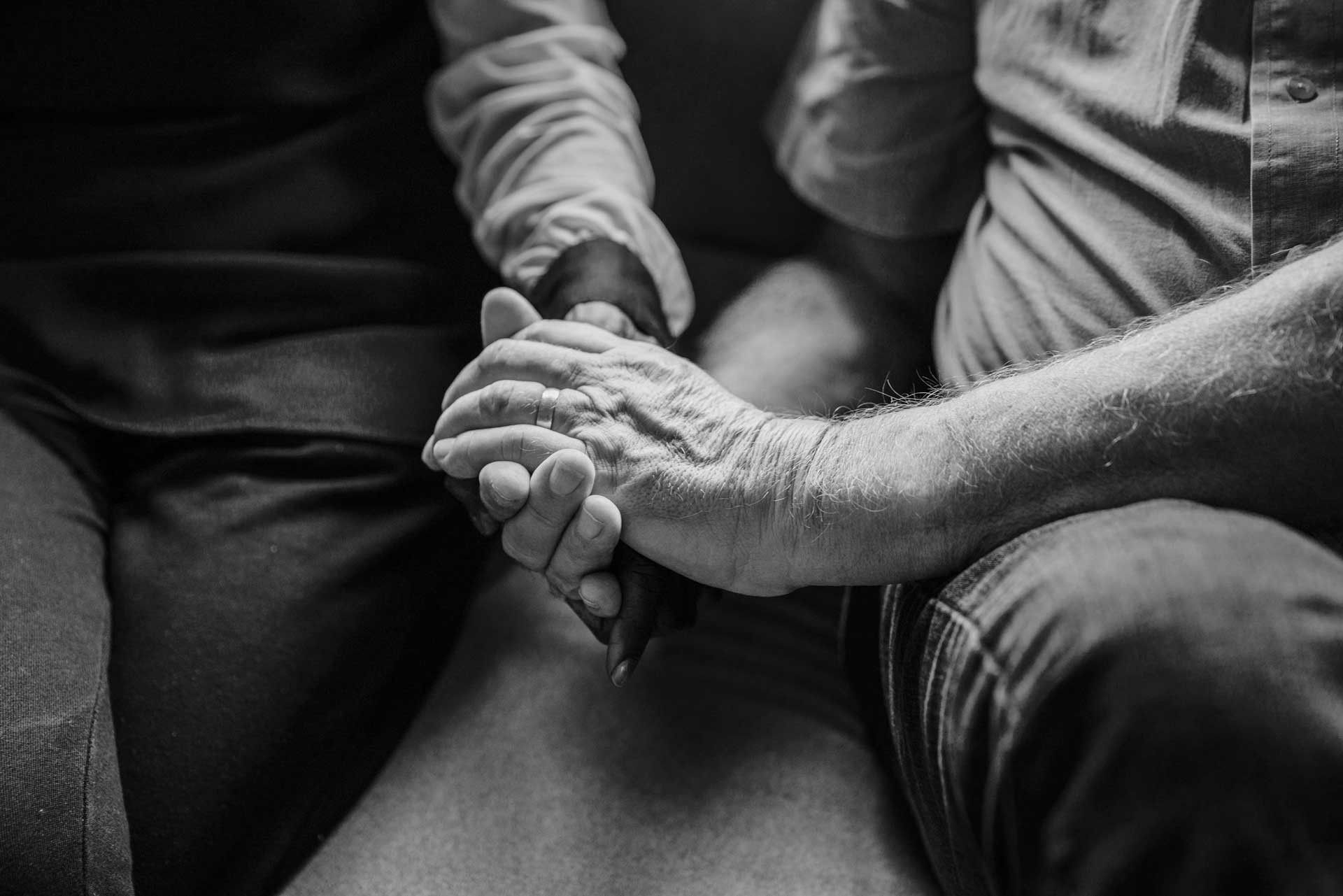 Black-and-white photo of elderly man wearing wedding band on ring finger holds someone else's hand with both hands